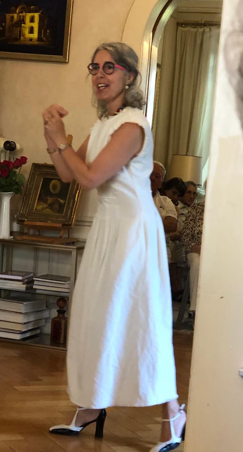 Elisabetta Aebischer-Gobbi en robe blanche et lunettes colorées, en pleine conférence devant un public assis dans un salon.